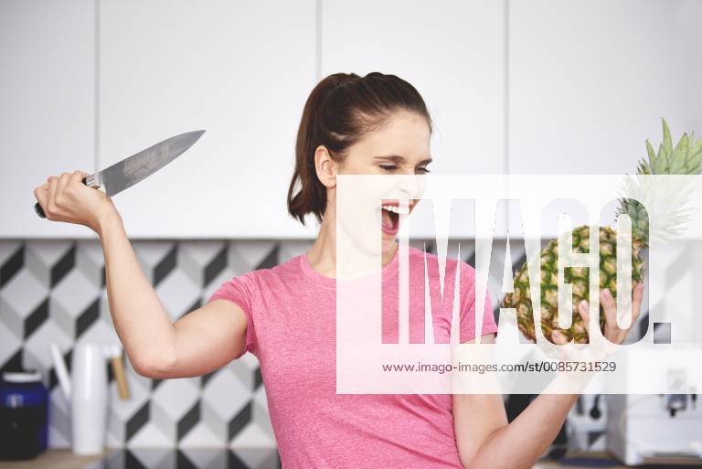 Portrait of screaming woman attacking pineapple with knife in the kitchen model released Symbolfoto property released ABIF00787