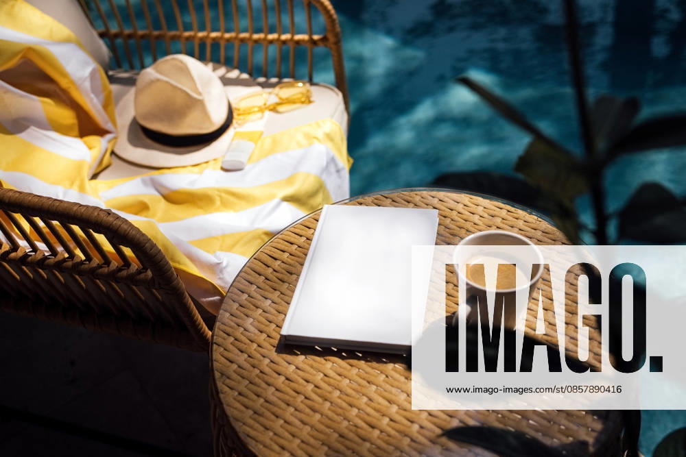 Gemütliche Atmosphäre im Freien mit einem Korbstuhl, auf dem eine gelb gestreifte Decke liegt, einem Sonnenhut, einem leeren Notizbuch und einer Tasse Kaffee am Rand eines Swimmingpools Outdoor scene with a wicker chair adorned with a yellow striped blanket, a sun hat, a blank notebook, and a cup of coffee next to a swimming pool (License=RF)