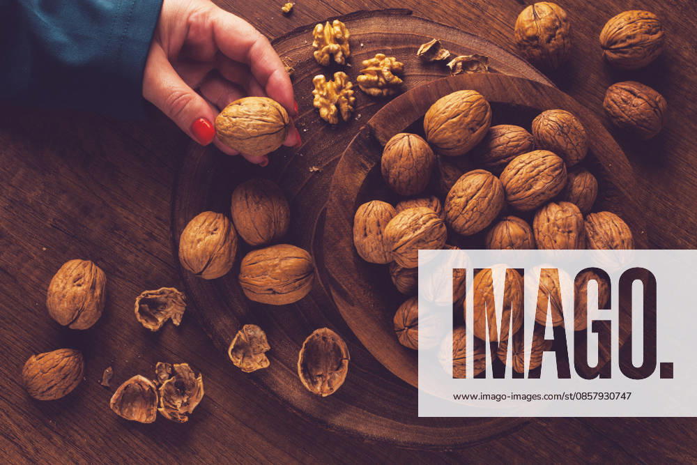 Die Hand einer Frau, die einen Walnuss aus einer Holzschale mit Walnüssen nimmt. Es gibt Schalen und Walnüsse, die herum verstreut sind. a woman s hand taking a walnut from a wooden bowl filled with walnuts. There are shells and walnuts scattered around. (License=RF)