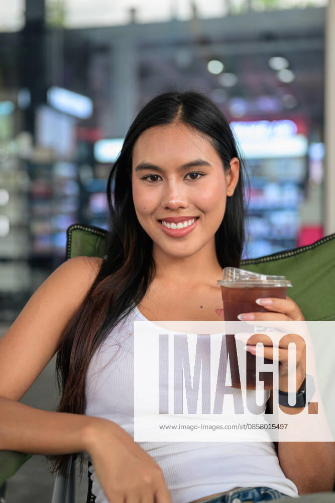 IMAGO Creative, Food, Trend, Iced Coffee Eine lächelnde Thailänderin, die einen Plastikbecher mit Eiskaffee in der Hand hält und im Freien in die Kamera blickt. Vertical medium close-up portrait of a cheerful young Isan Thai woman smiling and making direct eye contact with the camera. Seated relaxed in a green folding camping chair outdoors, she holds a clear plastic takeaway cup filled with refreshing iced coffee or cold brew against a softly blurred modern retail storefront background under bright daylight. (License=RF) ,model released, Symbolfoto