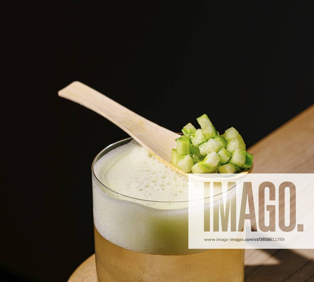 Food, Ein klares Getränk, das in einem Glas serviert wird, steht auf einer Holzoberfläche. Ein Holzlöffel mit fein gehacktem grünem Gemüse ruht auf dem Glas und verleiht dem Getränk eine frische Note Food, A clear beverage served in a glass sits on a wooden surface. A wooden spoon with finely chopped green vegetables rests on the glass, adding a fresh touch to the drink