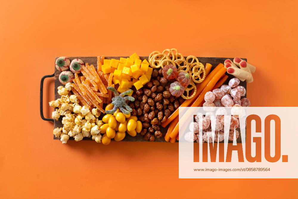 Halloween-Wurst- und Käseplatte mit Kürbis, Schokoladenbonbons, Spinne und Crackern. Halloween sweet charcuterie board with pumpkin, popcorn, dried fruits, color chocolate candy, spider, crackers on orange background. Festive food for kids party. View from above. (License=RF)