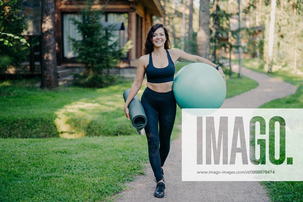 Eine lächelnde Frau trägt einen Gymnastikball und eine Trainingsmatte und geht auf einem Weg in der Nähe eines Holzhauses im Wald spazieren Smiling woman carries fitness ball and exercise mat walking along path near wooden house in forest (License=RF) ,model released, Symbolfoto
