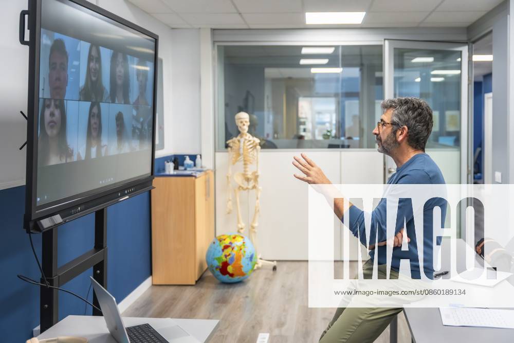 Der Lehrer leitet den virtuellen Anatomieunterricht von einem Labor aus und erklärt Skelettstrukturen neben einem menschlichen Skelett, während er mit den Schülern über eine Großbildvideokonferenz für E Learning interagiert Teacher leads virtual anatomy class from a lab, explaining skeletal structures beside a human skeleton while interacting with students via large screen video conference for e learning