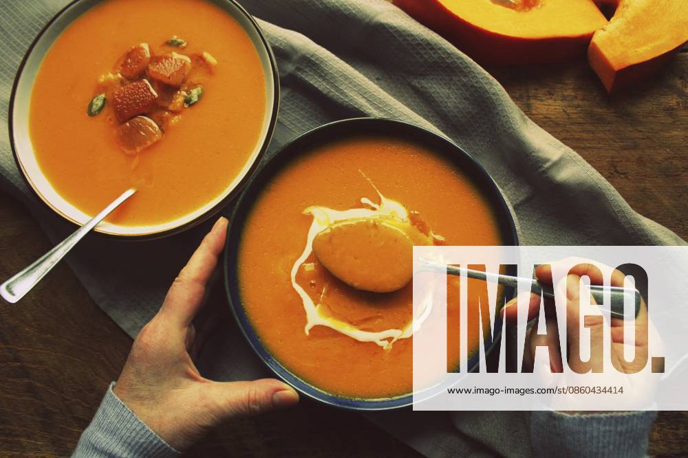 Flache Frauenhände und herbstwärmende Kürbiscremesuppe mit Croutons und Samen an Bord auf rustikalem Holzgrund, Ansicht von oben. Vegetarisches, veganes, gesundes Hausmannskostkonzept im Herbst Flat-lay of female hands and Fall warming pumpkin cream soup with croutons and seeds on board over rustic wooden background, top view. Autumn vegetarian, vegan, healthy comfort food eating concept