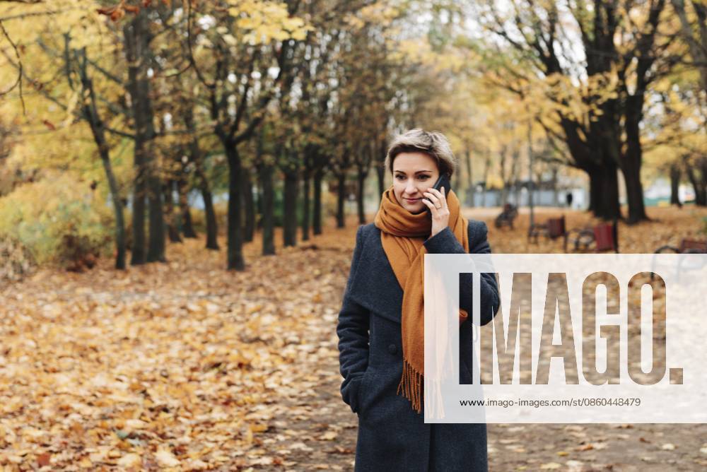 Kurzhaarige blonde Frau kommuniziert am Telefon und geht in Mantel und Schal im Herbstpark spazieren Short-haired blonde woman communicates on the phone walking in the autumn park in a coat and scarf