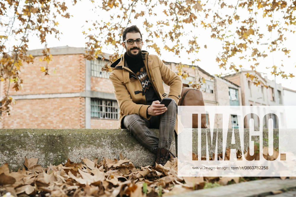 Spain, Igualada, portrait of smiling man with cell phone sitting down in the autumnal town model released, Symbolfoto, JRFF02297
