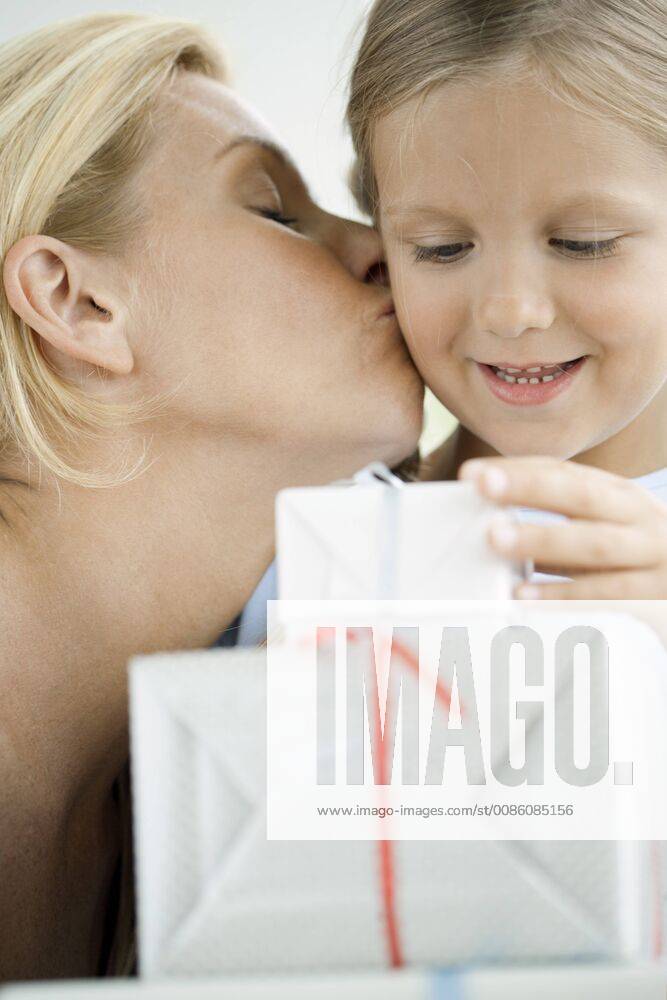 Mother kissing young daughter s cheek as girl looks at wrapped gifts