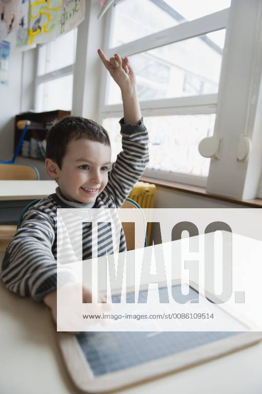 Boy sitting at desk with chalkboard, hand raised