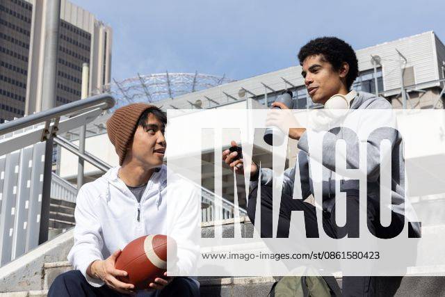 Diverse male friends sitting on concrete stairs wearing hoodies holding football and water bottle, model released, 08.08.2025 06:00:00, 7489140