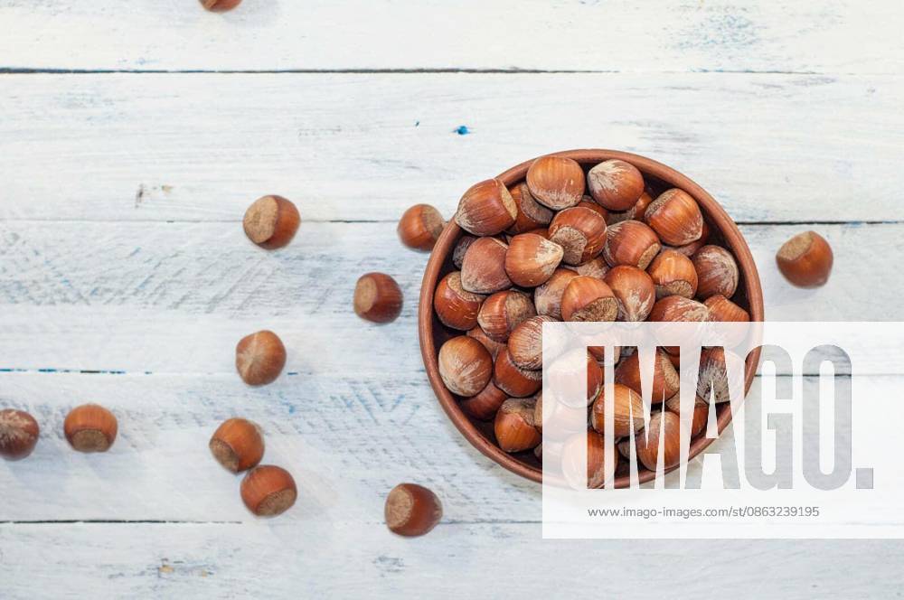 Hazelnuts in shell in brown clay bowl on a white wooden background, top view