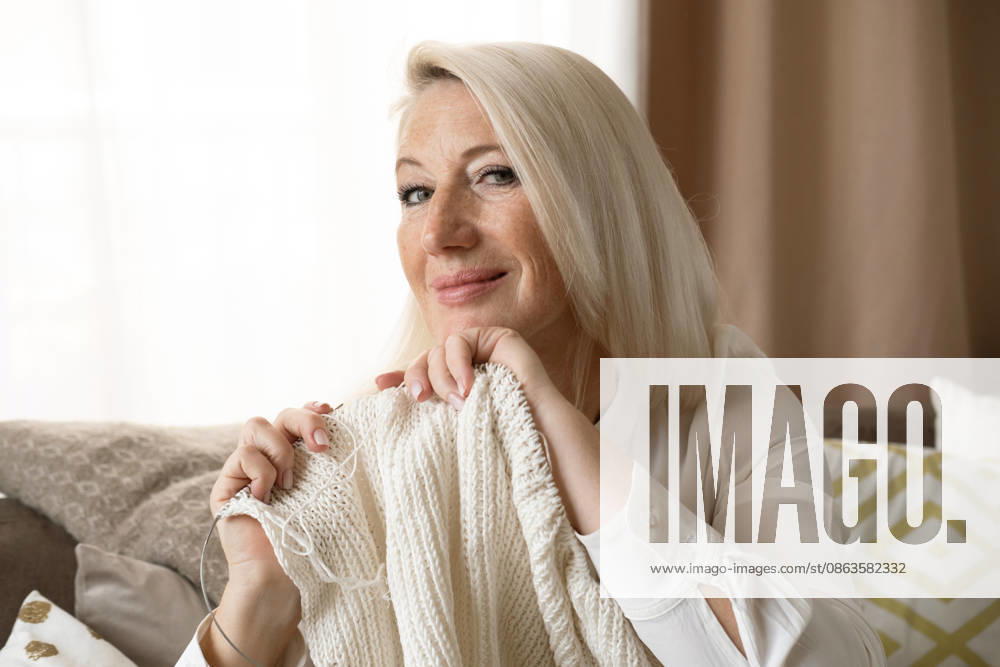 Eine ältere blonde Frau sitzt zu Hause auf dem Sofa, hält Stricknadeln und Wolle in der Hand und strickt Kleidung für ihre Lieben. Eine ältere blonde Frau sitzt zu Hause auf dem Sofa, hält Stricknadeln und Wolle in der Hand und strickt Kleidung für ihre Lieben. (License=RF) ,model released, Symbolfoto