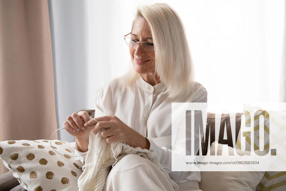 Eine ältere blonde Frau sitzt zu Hause auf dem Sofa, hält Stricknadeln und Wolle in der Hand und strickt Kleidung für ihre Lieben. Eine ältere blonde Frau sitzt zu Hause auf dem Sofa, hält Stricknadeln und Wolle in der Hand und strickt Kleidung für ihre Lieben. (License=RF) ,model released, Symbolfoto