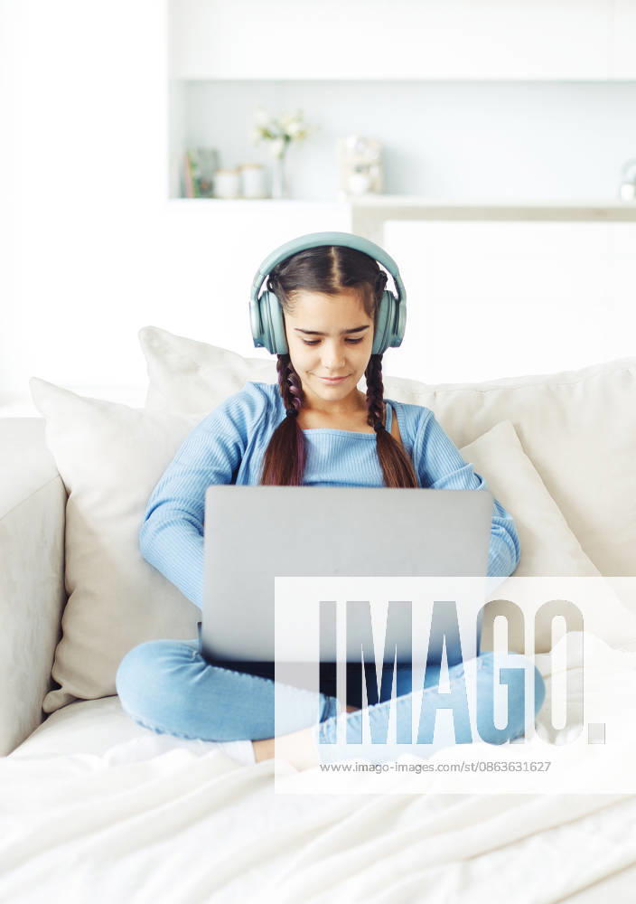 Ein Schulmädchen mit Kopfhörern sitzt auf dem Sofa und arbeitet an einem Laptop Ein Schulmädchen mit Kopfhörern sitzt auf dem Sofa und arbeitet an einem Laptop (License=RF) ,model released, Symbolfoto