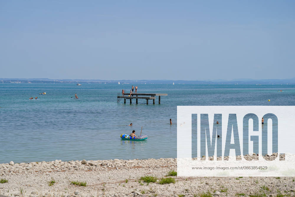 Lake Constance: Swimmers and water sports enthusiasts enjoy a sunny summer day on Lake Constance. People are swimming in the lake, riding in inflatable boats, and stand-up paddleboarding; others are standing on a swimming dock. In the background are sailboats and the opposite shore. Summer, heat, leisure, swimming, water sports, touri