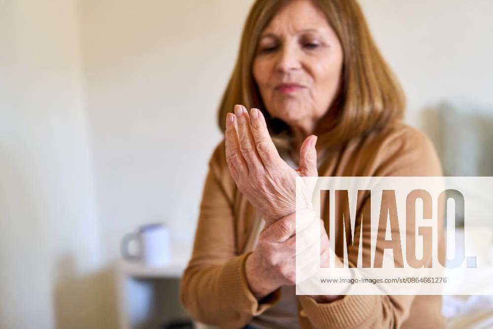 Senior woman experiencing wrist pain, home healthcare and elderly care concept, aging and joint discomfort. Older adult female holding her wrist in a bright, minimalist home interior, suggesting a South American household. Focus on the hand and wrist, with soft focus on the woman s face, a sense of discomfort and the need for care. (License=RF) ,model released, Symbolfoto