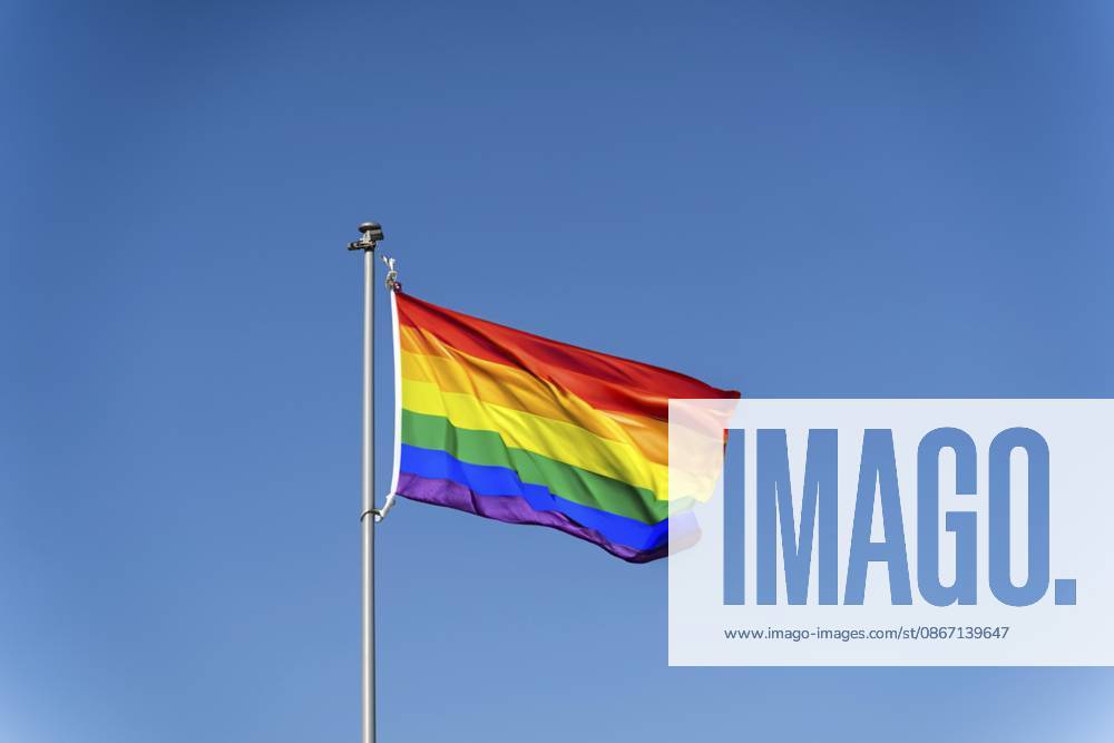 Die Regenbogenflagge der LGBTQ Community weht vor dem Hintergrund eines blauen, klaren Himmels. Eine Regenbogenflagge weht vor einem klaren blauen Himmel und symbolisiert den LGBTQ Stolz The rainbow flag of the LGBTQ community flies against the backdrop of a blue clear sky, A rainbow flag waving against a clear blue sky symbolizing LGBTQ pride