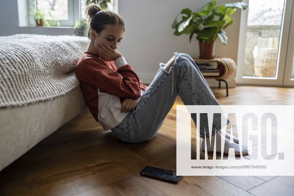 Das beleidigte unglückliche Teenager Mädchen schaut traurig auf das Telefon und sitzt zu Hause auf dem Boden. Trauriges, nachdenkliches, besorgtes Schulmädchen, das nach der Trennung von einem geliebten Jungen oder einem verletzenden Beitrag in den sozialen Medien an Depressionen leidet Offended unhappy teenage girl sadly looking at phone sits on floor at home. Sad thoughtful worried schoolgirl experiencing depression after breaking up with beloved boy or hurtful social media post