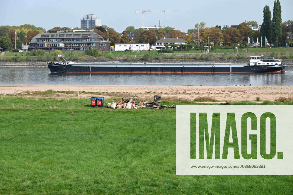 Low water levels in the Rhine have exposed a large sandbar; rocks and sand are visible on the riverbank near the Theodor Heuss Bridge; a cargo ship is sailing in the channel; in the foreground, flotsam, car tires, and debris have been exposed by the low water, Unidentified individuals collected the trash and left it on the bank for pickup; low water levels