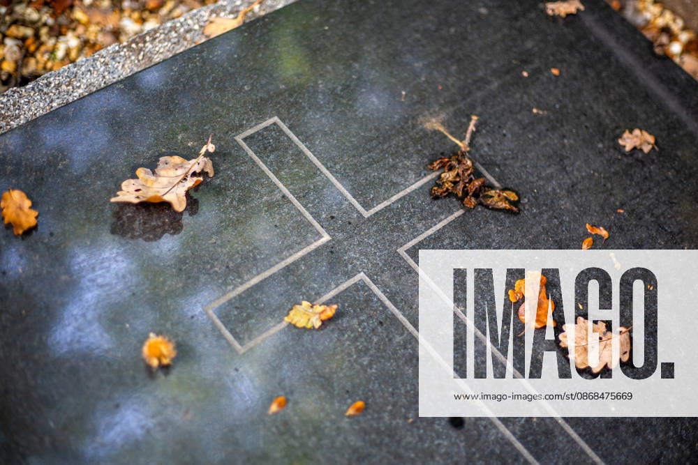 On a dark, polished gravestone with an engraved cross, a few brown oak leaves lie in the autumn light all around Eicks