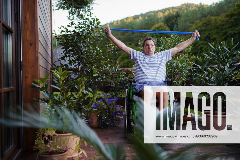 Senior man in wheelchair exercising with resistance band on terrace. Senior man with physical disability exercising independently on terrace. model released
