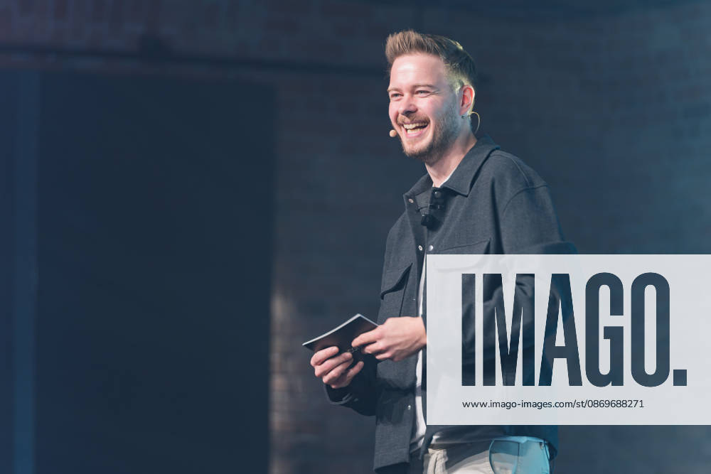 Sascha Müller, host of NeulandPro Connect 2026, stands on stage in front of a dark brick wall, laughing with his cue ca