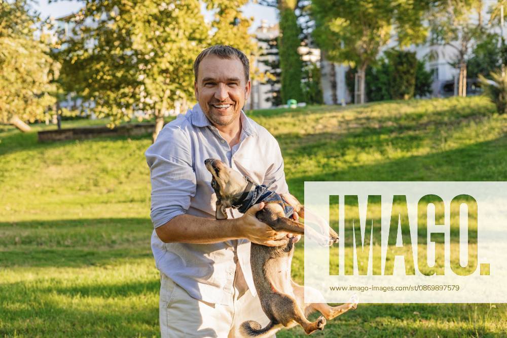 Ein lächelnder Mann wiegt seinen kleinen Hund in einem sonnigen Park A smiling man cradles his small dog in a sunny park