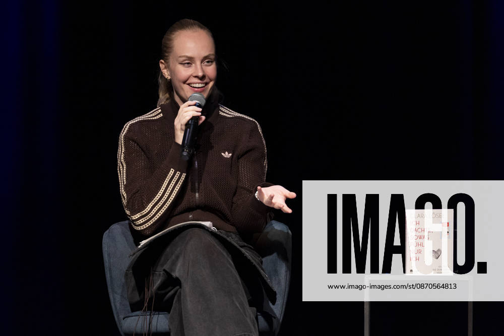 Poetry artist and author Clara LOESEL LÖSEL speaks to the audience with a smile during her reading—a preview show for her new book, *Ich dachte, sowas fühle nur ich*—seated with a microphone, in a single frame, a single photo, at the Theater am Tanzbrunnen, Cologne, on October 23, 2026