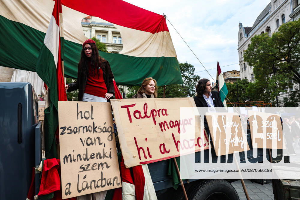 Young participants dressed in period clothing stand aboard a vintage truck beneath a Hungarian flag with its communist emblem removed during a historical reenactment commemorating the 70th anniversary of the 1956 Hungarian Revolution. The participants display signs bearing historical slogans, including Talpra magyar, hi a haza! (Rise up, Hungarian, the homeland calls!), A feny a feny (The light is the light), A szel a szel (The wind is the wind), and Hol zsarnoksag van, mindenki szem a lancban (Where there is tyranny, everyone is a link in the chain). The flag with its centre cut out became a symbol of the uprising against Soviet-backed rule. Budapest Hungary - ZUMAo116 20261023_znp_o116_012