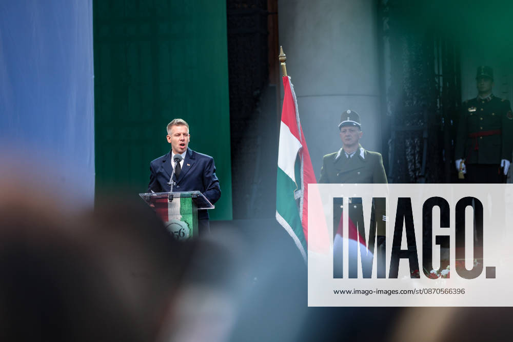 Seen through a blurred crowd, Hungarian Prime Minister PETER MAGYAR speaks at a podium beside a ceremonial military guard and a Hungarian revolutionary flag with its centre cut out, during commemorations marking the 70th anniversary of the 1956 Hungarian Revolution outside Parliament. Budapest Hungary - ZUMAo116 20261023_znp_o116_042