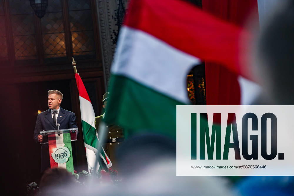 A large Hungarian revolutionary flag, with its centre cut out, waves in the foreground as Prime Minister PETER MAGYAR delivers his first October 23 commemorative address as prime minister outside Parliament, with a ceremonial military guard standing beside him during the 70th anniversary of the 1956 Hungarian Revolution. Budapest Hungary - ZUMAo116 20261023_znp_o116_058