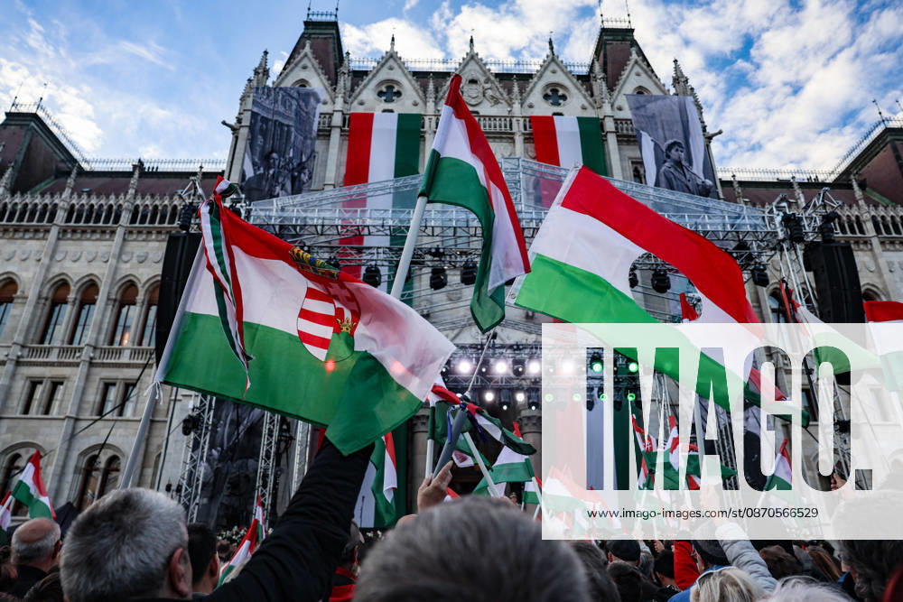 Hungarian flags, including the 1956 revolutionary flag with its centre cut out, wave above crowds gathered outside Parliament during commemorations marking the 70th anniversary of the Hungarian Revolution. Large historical photographs and Hungarian tricolours adorn the Parliament facade above the stage. Budapest Hungary - ZUMAo116 20261023_znp_o116_075