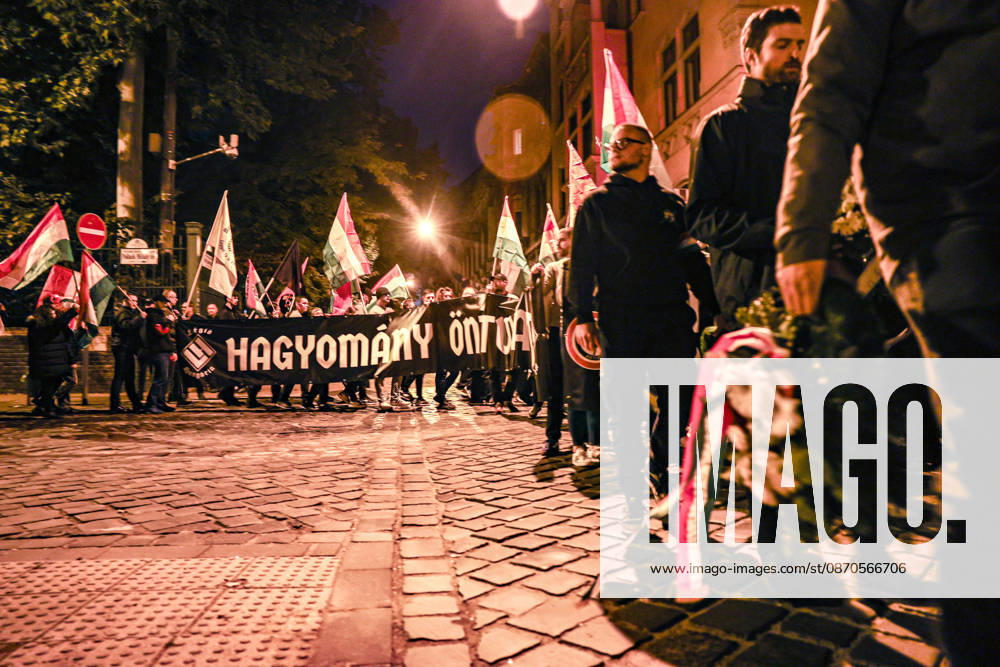Members of Legio Hungaria, a Hungarian far-right ultranationalist group, march along a cobblestone street behind a large black banner reading Hagyomany, Ontudat, Kozosseg (Tradition, Self-Awareness, Community), while waving Hungarian flags during a nighttime procession commemorating the 70th anniversary of the 1956 Hungarian Revolution and the 20th anniversary of the 2006 anti-government unrest. Budapest Hungary - ZUMAo116 20261023_znp_o116_095