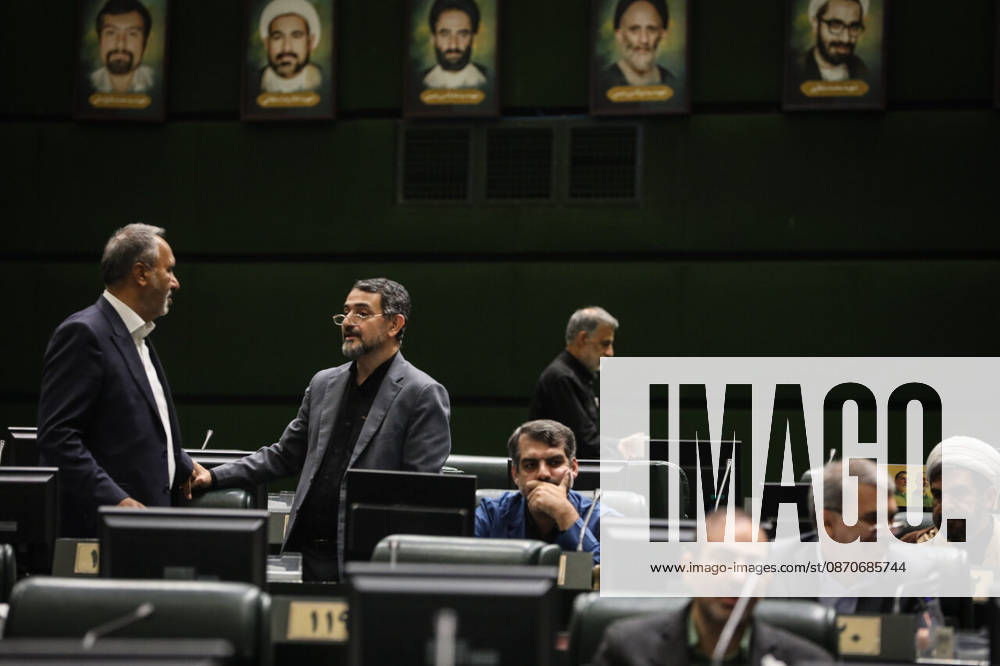 Two suited lawmakers stand talking beneath framed portraits as other members sit at their desks in the chamber at the Islamic Consultative Assembly in Tehran, Iran, on October 25, 2026. The Islamic Consultative Assembly resumed an open sitting in its Tehran chamber with a mix of in-person and remote participation, according to parliamentary reporting. Tehran Iran - ZUMAi98_ 20261025_zih_i98_045