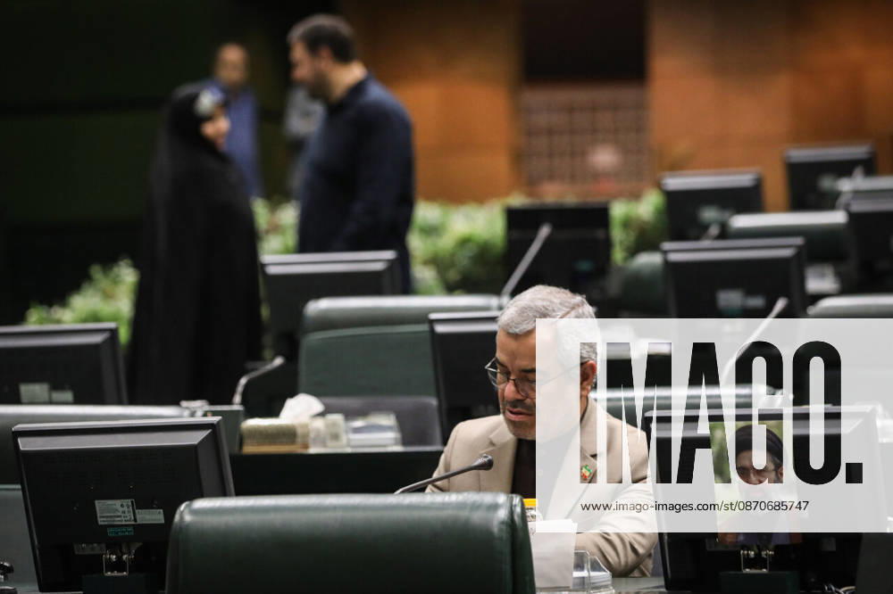 A lawmaker in a beige jacket looks down at his parliamentary desk as a woman in a black chador talks to a man behind him at the Islamic Consultative Assembly in Tehran, Iran, on October 25, 2026. The Islamic Consultative Assembly resumed an open sitting in its Tehran chamber with a mix of in-person and remote participation, according to parliamentary reporting. Tehran Iran - ZUMAi98_ 20261025_zih_i98_046