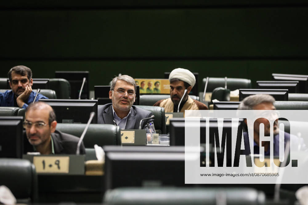 Several seated male lawmakers and a white-turbaned cleric occupy rows of green benches with digital desk displays at the Islamic Consultative Assembly in Tehran, Iran, on October 25, 2026. The Iranian parliament held a hybrid open sitting, with some lawmakers attending in the Tehran chamber and others participating remotely, according to parliamentary reports. Tehran Iran - ZUMAi98_ 20261025_zih_i98_051