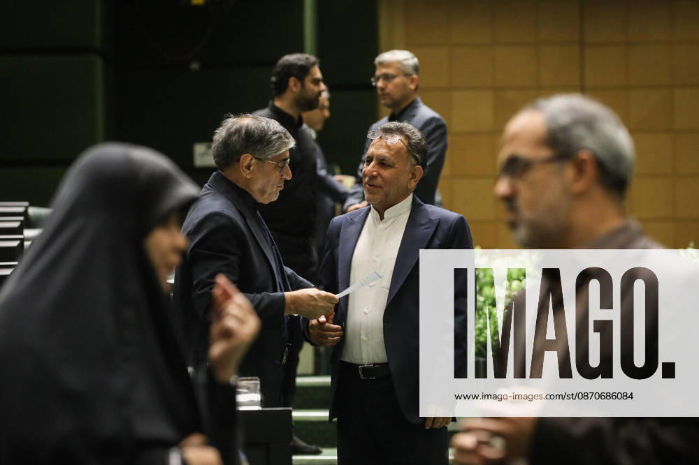 A suited lawmaker reaches to shake hands with a colleague while another man watches in the foreground at the Islamic Consultative Assembly in Tehran, Iran, on October 25, 2026. The Iranian parliament held a hybrid open sitting, with some lawmakers attending in the Tehran chamber and others participating remotely, according to parliamentary reports. Tehran Iran - ZUMAi98_ 20261025_zih_i98_083