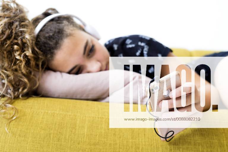 Girl lying on the couch at home listening music with headphones and smartphone model released Symbolfoto property released ERRF00053