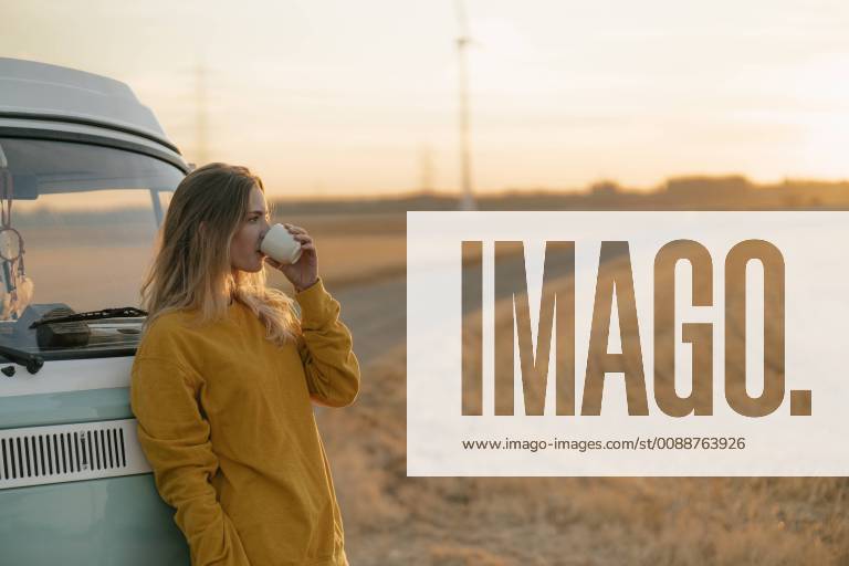 Young woman drinking from mug at camper van in rural landscape at sunset model released Symbolfoto GUSF01634