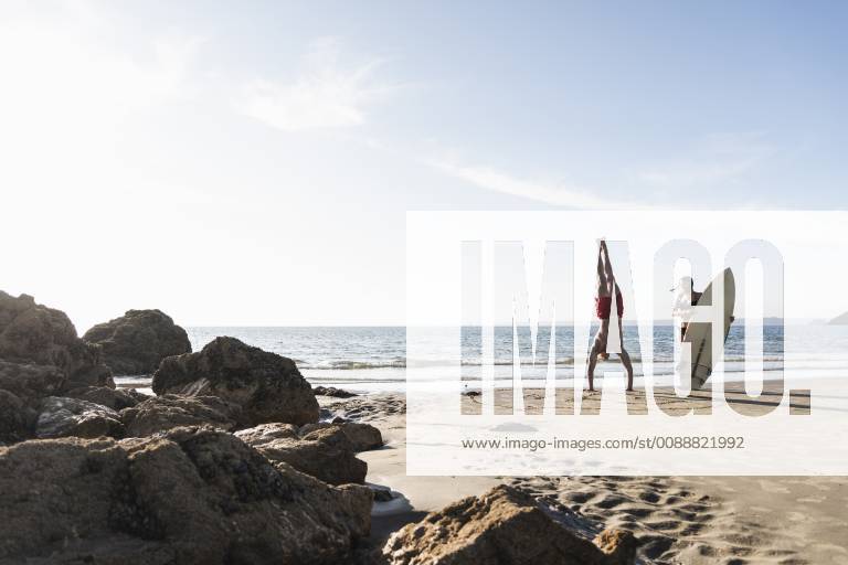 France, Brittany, young man doing a handstand and woman holding surfboard on the beach model released Symbolfoto UUF15913