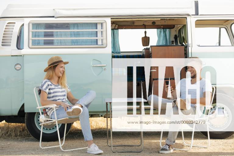 Happy young couple sitting on camping chairs at camper van with cell phone model released Symbolfoto GUSF01577