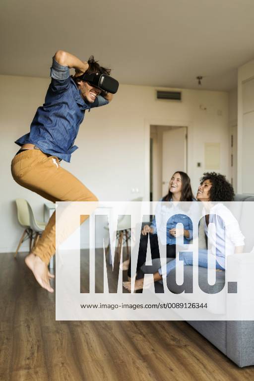 IMAGO Creative, Lifestyle, Gaming Two women watching man with VR glasses jumping at home model released Symbolfoto property released VABF01792