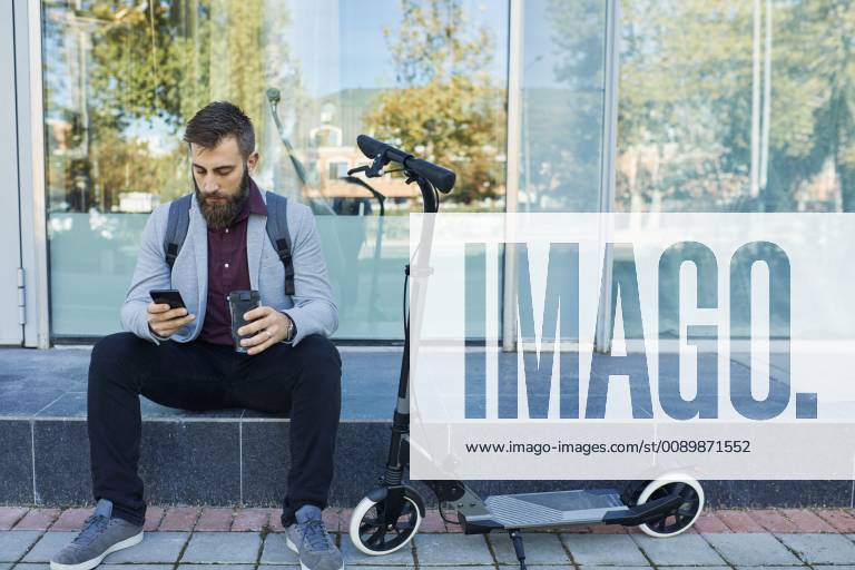 Businessman sitting outdoors using cell phone next to scooter model released Symbolfoto ZEDF01702