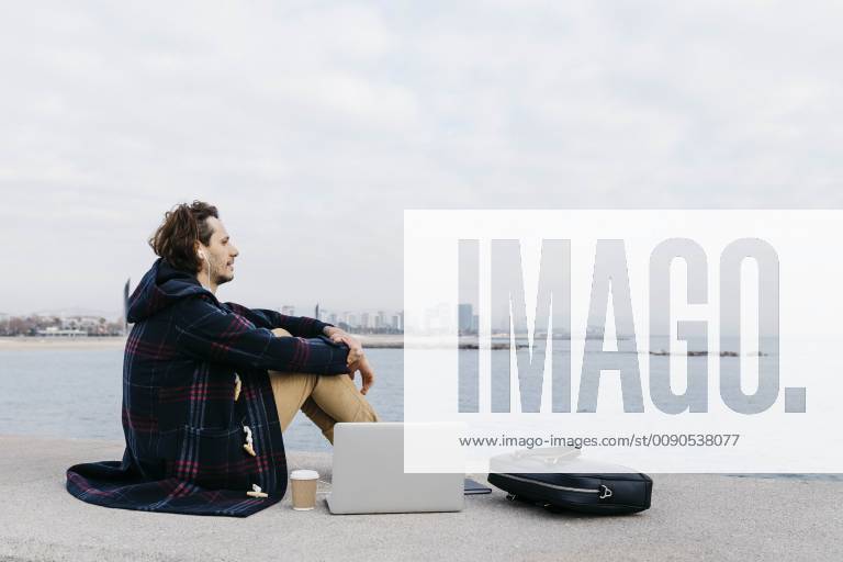 Spain, Barcelona, man with laptop, briefcase and earphones sitting at the sea model released Symbolfoto JRFF02516