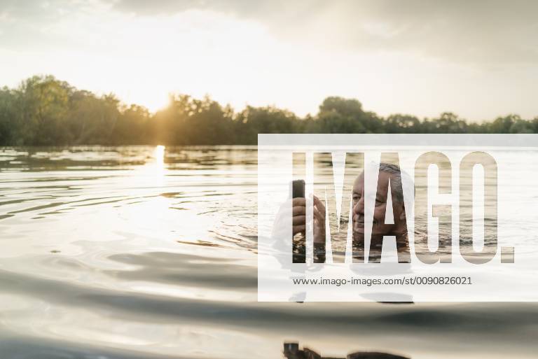 Senior man swimming in a lake at sunset using cell phone model released Symbolfoto GUSF01823