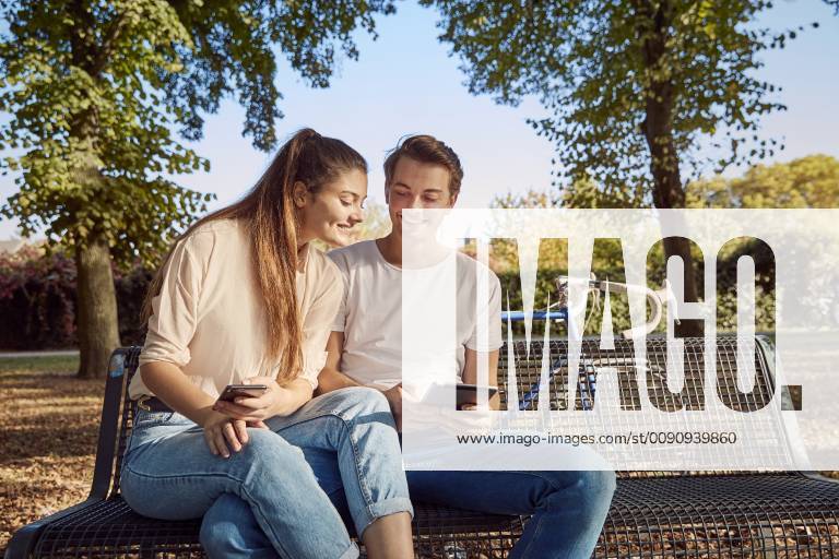 Young couple in park sitting on a bench with cell phone and tablet model released Symbolfoto JHAF00041