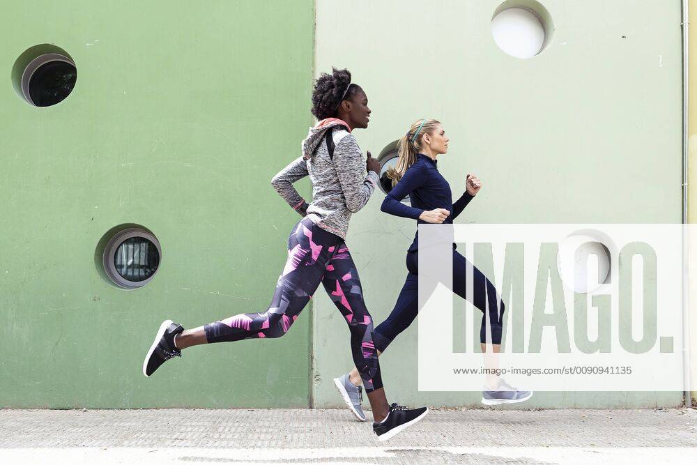 Two sporty young women running together in the city passing wall with round windows model released Symbolfoto JSRF00139
