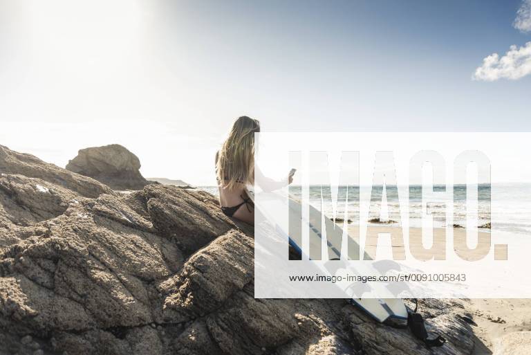 Young woman with surfboard sitting on a rock at the beach, using smartphone model released Symbolfoto UUF16485
