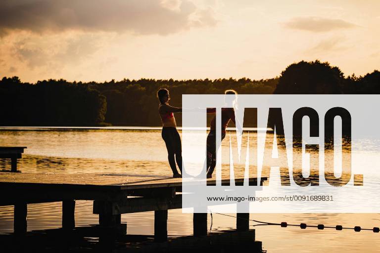 Women practicing acro yoga on tranquil sunset lake dock Germany Mecklenburg-Vorpommern Barnin