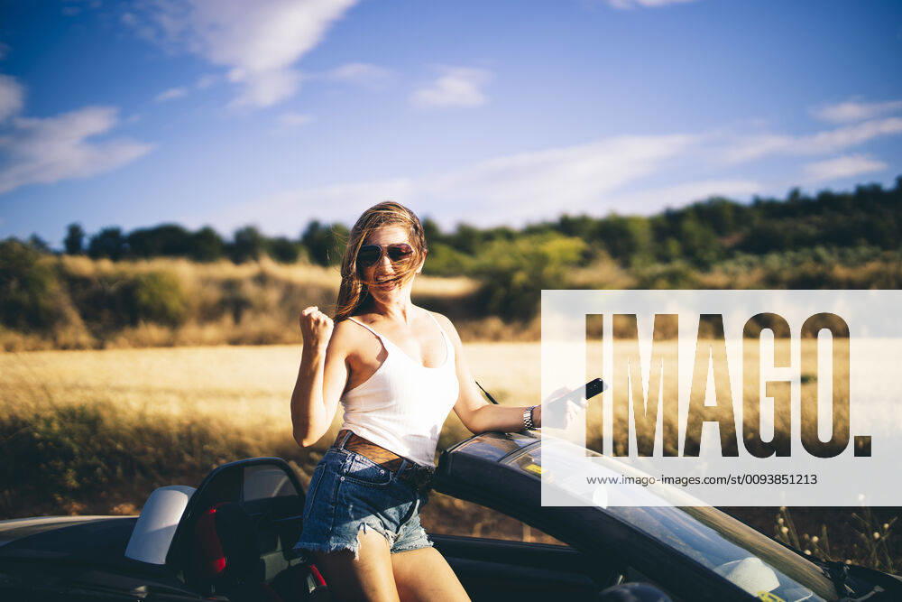Woman standing in her convertible, holding smartphone, happy about good news model released Symbolfoto OCMF00610